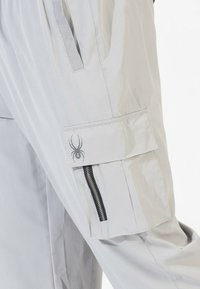 Light gray cargo pants feature a side pocket with a flap, zip closure, and a spider logo. The fabric appears smooth and lightweight.