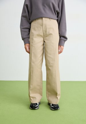 Pantaloni beige a gamba larga in cotone, con una texture liscia, pieghe frontali e un trattamento in vita regolare. Abbinati a scarpe sportive nere.