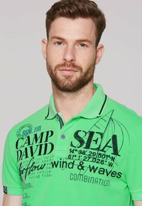 Zielona bawełniana koszulka polo z nadrukowanym tekstem i morskimi grafikami. Posiada kołnierzyk z zapięciem na guziki oraz kontrastowe wykończenie.