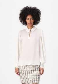 Blusa crema con cuello y detalle de ojal, con mangas plisadas y puños elásticos, combinada con una falda texturizada y multicolor.