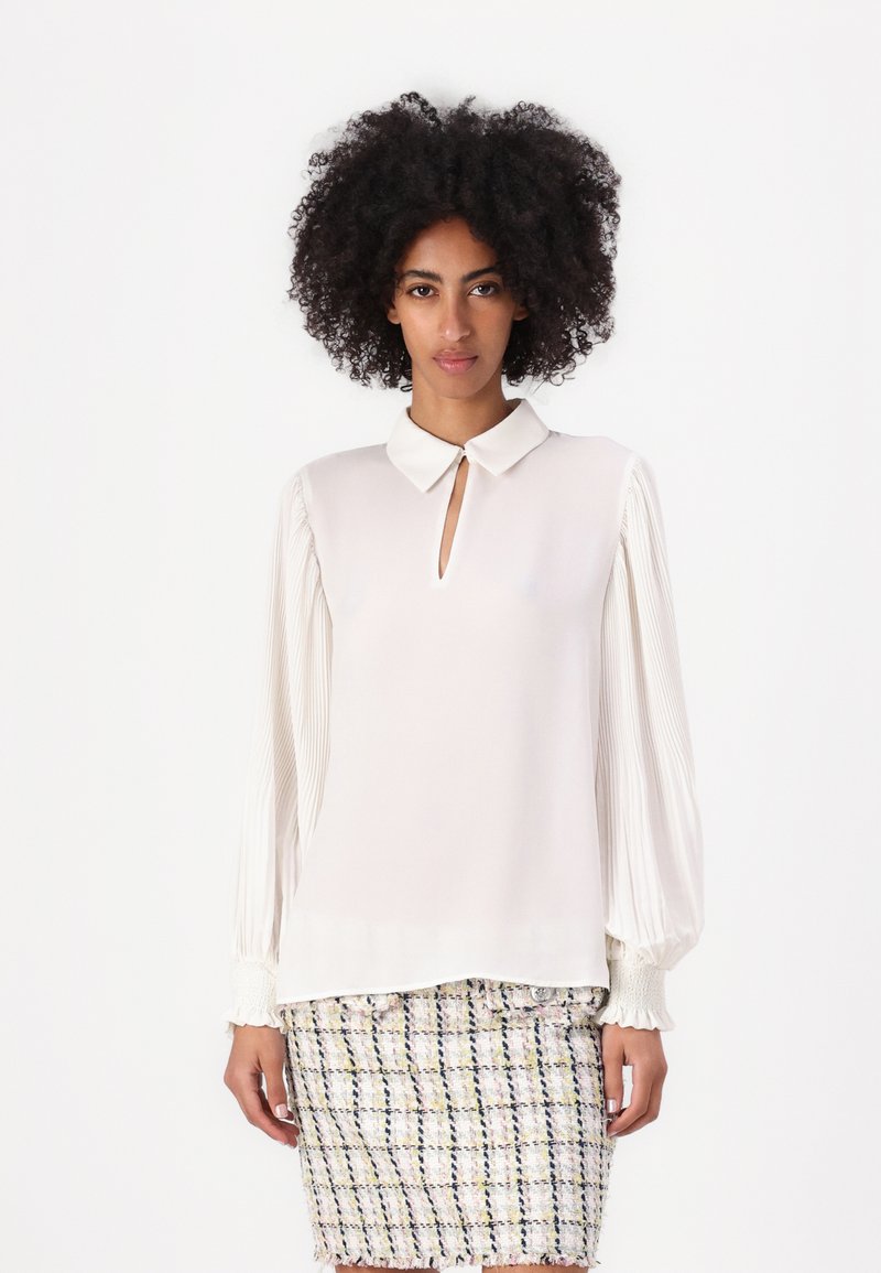 Blusa crema con cuello y detalle de ojal, con mangas plisadas y puños elásticos, combinada con una falda texturizada y multicolor.