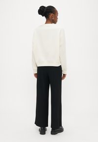 Hellbeiges Sweatshirt mit überschnittenen Schultern, geripptem Saum und weiten, schwarzen, gerippten Hosen. Das Modell trägt klobige schwarze Schuhe, von hinten gesehen.