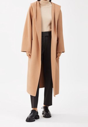Femme portant un long manteau camel, un pull à col roulé côtelé beige, un pantalon noir sur mesure et des chaussures à plateforme lacées noires, debout sur fond blanc.