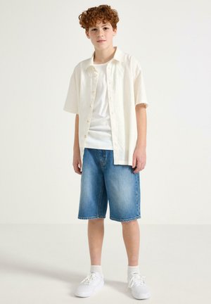 Rødharret dreng stående, iført hvid kortærmet skjorte med knapper over hvid T-shirt, knælange blå denimshorts, hvide sokker og hvide sneakers.
