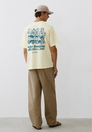 Uomo con cappellino beige, maglietta giallo chiaro con corridori dei cartoni animati blu e scritta sulla schiena, pantaloni marroni larghi e sandali neri, in piedi contro un muro bianco.