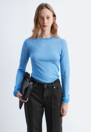 Jonge vrouw met een lichtblauw shirt met lange mouwen en zwarte broek, houdt een zwarte handtas vast met een patroon-sjaal, staand tegen een effen achtergrond.