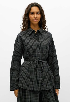 Un modèle aux longs cheveux foncés porte une robe-chemise à carreaux couleur anthracite, nouée à la taille, sur un fond blanc uni.