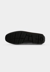 Semelle de chaussure noire avec des motifs de semelle profonds et géométriques, vue de dessous sur un fond gris clair.