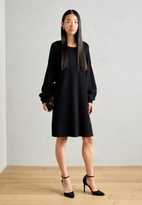 Black knit dress with long, puffed sleeves and a round neckline. Model paired with black ankle-strap heels and a small black handbag.