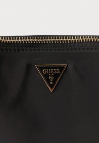 Μαύρο τσαντάκι χειρός από νάιλον με φερμουάρ σε χρυσή απόχρωση και τριγωνική μαύρη και χρυσή πινακίδα με το λογότυπο "GUESS" και ένα ερωτηματικό.