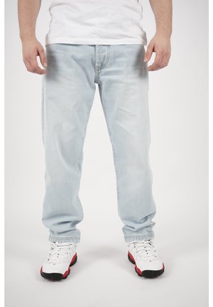 Mann trägt hellblaue Jeans, weiße Sneakers mit roten und schwarzen Akzenten, steht vor einem schlichten Hintergrund.