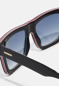 Occhiali da sole neri con lenti blu sfumate, caratterizzati da un audace design geometrico e da dettagli a strisce rosse e bianche lungo il bordo superiore della montatura.