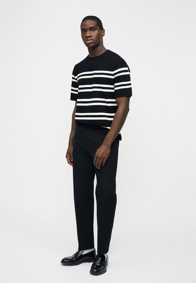 Άνδρας που στέκεται φορώντας μαύρο πουλόβερ με κοντά μανίκια και λευκές ρίγες, μαύρο παντελόνι, λευκές κάλτσες και μαύρα μοκασίνια, μπροστά σε απλό φόντο.