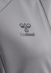 Grå sportjacka med texturerad yta, med en mörkgrå hummel-logotyp på vänster bröst. Dragkedja är synlig.