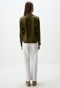 Chemise à manches longues en satin vert olive avec col, associée à un pantalon blanc à jambe droite et des chaussures à enfiler beiges. Texture lisse, détails minimaux.