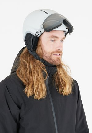 Witte sk helm met vizier en zwarte skibril, gedragen met een zwarte jas. Heeft lang bruin haar en een lichte baard.