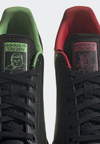 Czarne sneakersy adidas Stan Smith mają podwójne języki w dwóch kolorach: jeden zielony z graficznym wzorem, drugi czerwony, oba z czarnymi sznurowadłami.