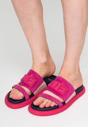 Piedi che indossano sandali aperti rosa e blu navy con due cinturini, uno largo con lettere in rilievo su sfondo grigio.