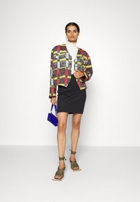 Mehrfarbige gemusterte Jacke, weißer Rollkragenpullover, schwarzer Rock, grüne geflochtene Sandalen und blaue Handtasche. Glattes Material und strukturierte Gestaltung.