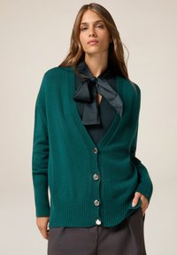 Cardigan teal realizzato in materiale morbido, con scollatura a V, polsini e orlo a coste, e bottoni argentati sul davanti. Senza motivi o disegni.