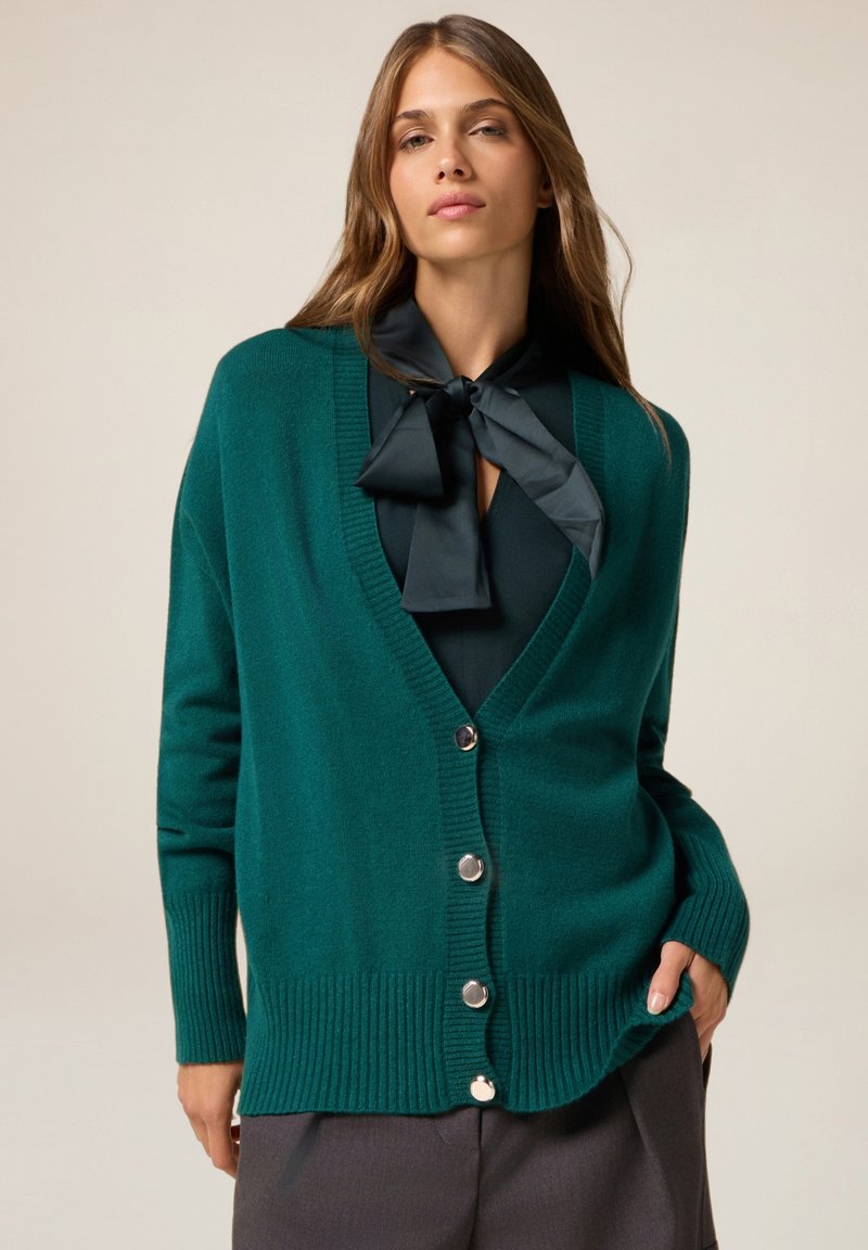 Cardigan teal realizzato in materiale morbido, con scollatura a V, polsini e orlo a coste, e bottoni argentati sul davanti. Senza motivi o disegni.