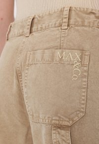 Beige fabric pants feature a back pocket with embossed "MAX&CO." logo. The texture is smooth with a subtle sheen.