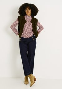 Jeans oscuros con detalles en botones dorados, combinados con una camisa de cuadros en color burdeos y un chaleco verde oliva. Botines de tacón en color beige completan el atuendo.