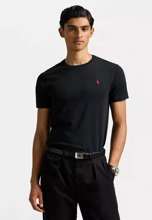 T-shirt nera a maniche corte in cotone, con un logo rosso sul lato sinistro del petto, abbinata a pantaloni neri e una cintura di pelle.