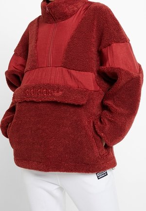 Person iført en rød Adidas fleece-pullover med lynlåsåbning ved kraven og kængurulomme, kombineret med hvide Adidas bukser.