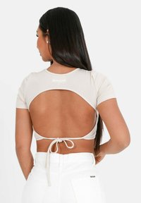 Crop top a coste color beige chiaro con maniche corte, design con schiena scoperta e chiusura a fiocco in vita. Nome del marchio "SIXTH JUNE" visibile nella parte superiore.