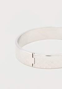 Ασημένιο βραχιόλι με χαρακτηριστική λεία, γυαλισμένη επιφάνεια και γεωμετρικό μοτίβο χαραγμένο στη μία πλευρά, με ασφαλή κλείσιμο με μεντεσέ.