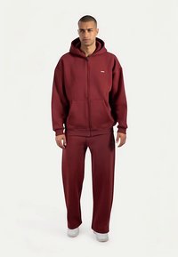 Sudadera granate con cremallera y bolsillos grandes, junto con pantalones de chándal de corte holgado a juego. Confeccionados en tela suave, presentan un diseño con una marca mínima y puños acanalados.