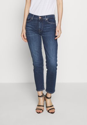 Femme portant un jean slim bleu foncé, un haut blanc et des sandales noires à talons hauts avec des brides, debout devant un fond uni.