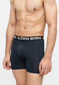 Άνδρας με γραμμωτό κορμό φορώντας μαύρα μπόξερ Björn Borg με λογότυπο στη μέση, στέκεται μπροστά σε ανοιχτό φόντο.