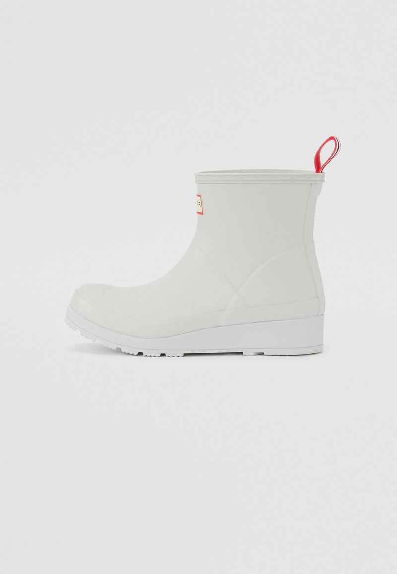 Botte de cheville en caoutchouc blanc imperméable avec surface texturée, semelle plate et une languette rouge à l'arrière. Présente un logo en patch sur le côté.