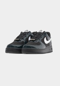 Μαύρα και σκούρα γκρι παπούτσια Nike Air Force 1 με λευκά λογότυπα Swoosh και μαύρα κορδόνια, στραμμένα προς τα αριστερά σε απλό λευκό φόντο.