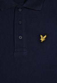 Marineblå polo-skjorte laget av teksturert stoff, med en tre-knappers glidelås og en brodert gul fugl-logo på venstre bryst.