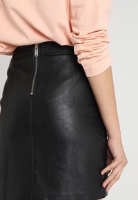 Hame hitam dari kulit sintetis dengan finishing tekstur dan ritsleting terlihat di bagian belakang. Dipadukan dengan atasan lengan panjang berwarna pink muda.