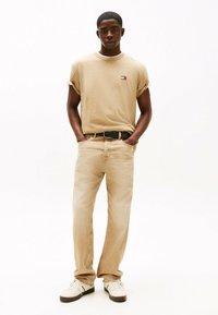 T-shirt color cammello con maniche arrotolate, dotata di un piccolo logo. Abbinata a jeans beige chiaro e sneakers bianche con dettagli neri.