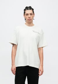 Biała bawełniana koszulka o luźnym kroju, z krótkimi rękawami i okrągłym dekoltem. Na górnej lewej stronie znajduje się nadrukowane czarne logo.