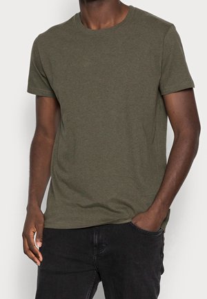 Man wearing plain olive green short-sleeve crew neck t-shirt with black jeans, one hand in pocket against a plain background.
