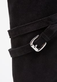 Botín de ante negro con una correa estrecha y detalle de hebilla de metal plateado, que muestra una textura suave y un diseño aerodinámico.