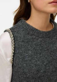 Sivi vuneni top bez rukava s okruglim izrezom; ima složene perlice duž šava na ramenu; mekana tekstura. Nošen preko bijele košulje.