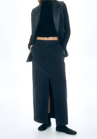 Jupe longue en jean foncé avec fente frontale, fermeture par boutons et poches latérales, associée à un top court noir et des chaussures plates noires.