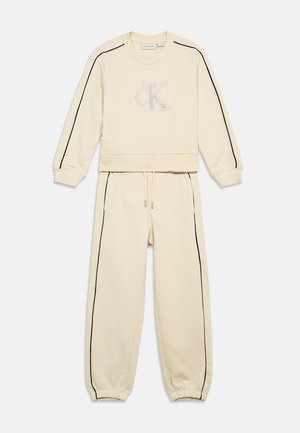 Conjunto de sudadera y pantalones deportivos color crema con rayas negras laterales y un sutil logo "CK" en el pecho, que cuenta con puños elásticos y pantalones con cordón ajustable.