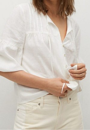 Kvinde iført en hvid skjorte med knapper og trekvartlange ærmer samt off-white bukser, der justerer skjortens nederste kant.