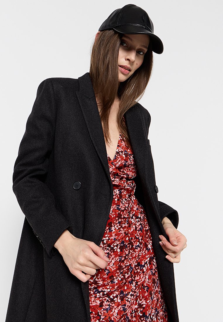 Manteau en mélange de laine noir, croisé avec des revers à notch, superposé sur une robe florale rouge. Le modèle porte une casquette noire.