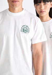 Weiße Baumwoll-T-Shirts mit grünen Drucken von Gemüse und dem Text "Green Chef Weekends" auf der linken Brustseite.