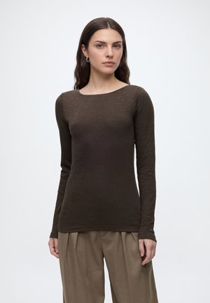 Vrouw met lang donker haar draagt een donkerbruine top met lange mouwen en een losse beige broek, staand tegen een effen lichte achtergrond.