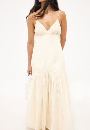 Frau trägt ein ärmelloses cremefarbenes Maxikleid mit dünnen Trägern und einem gerafften Rock, hält eine weiße strukturierte Handtasche.